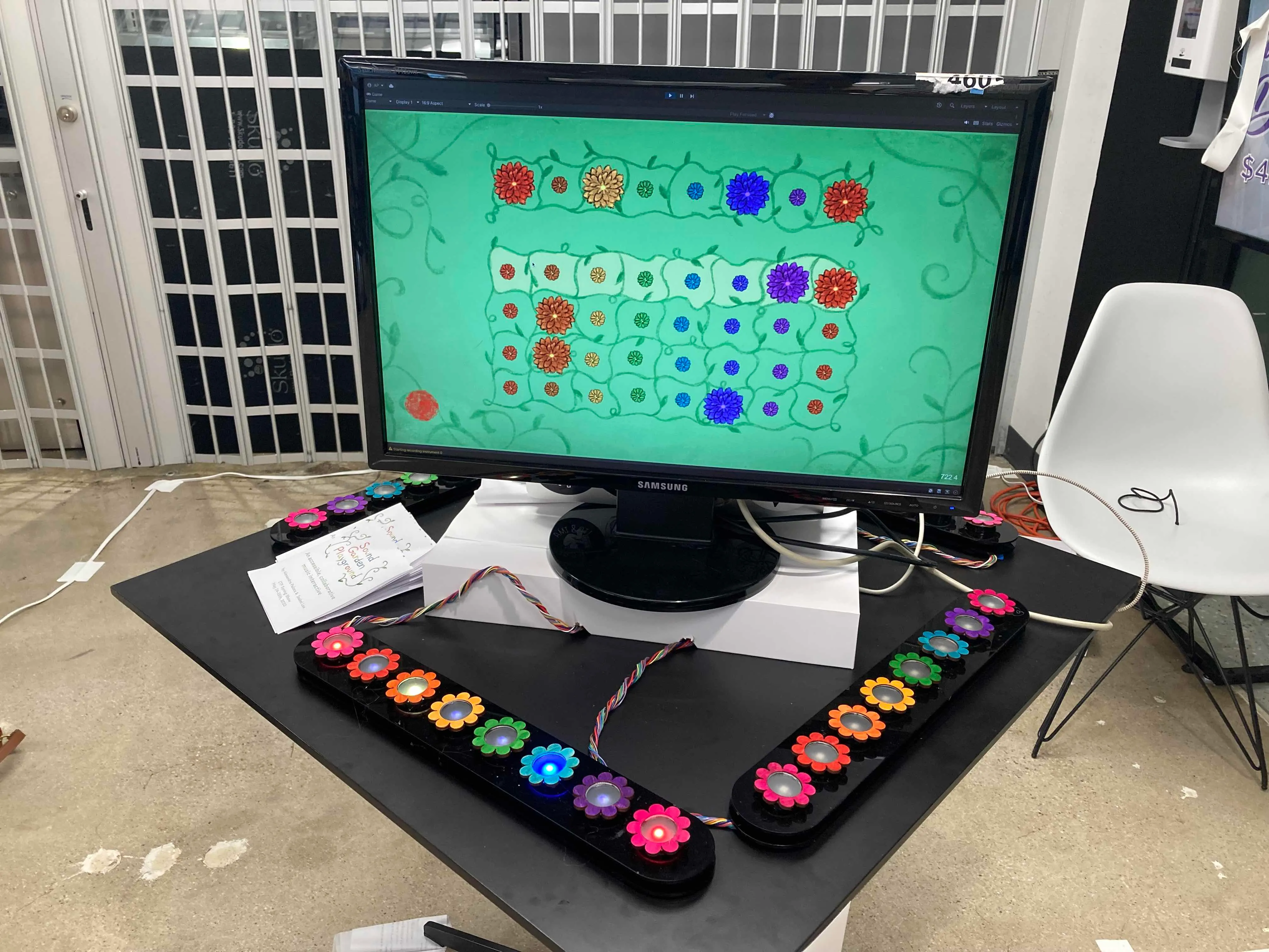 A screen in the center of a table shows a visual display of colorful flowers in rows. 
        Below the screen are rows of flower-shaped buttons with the same colors