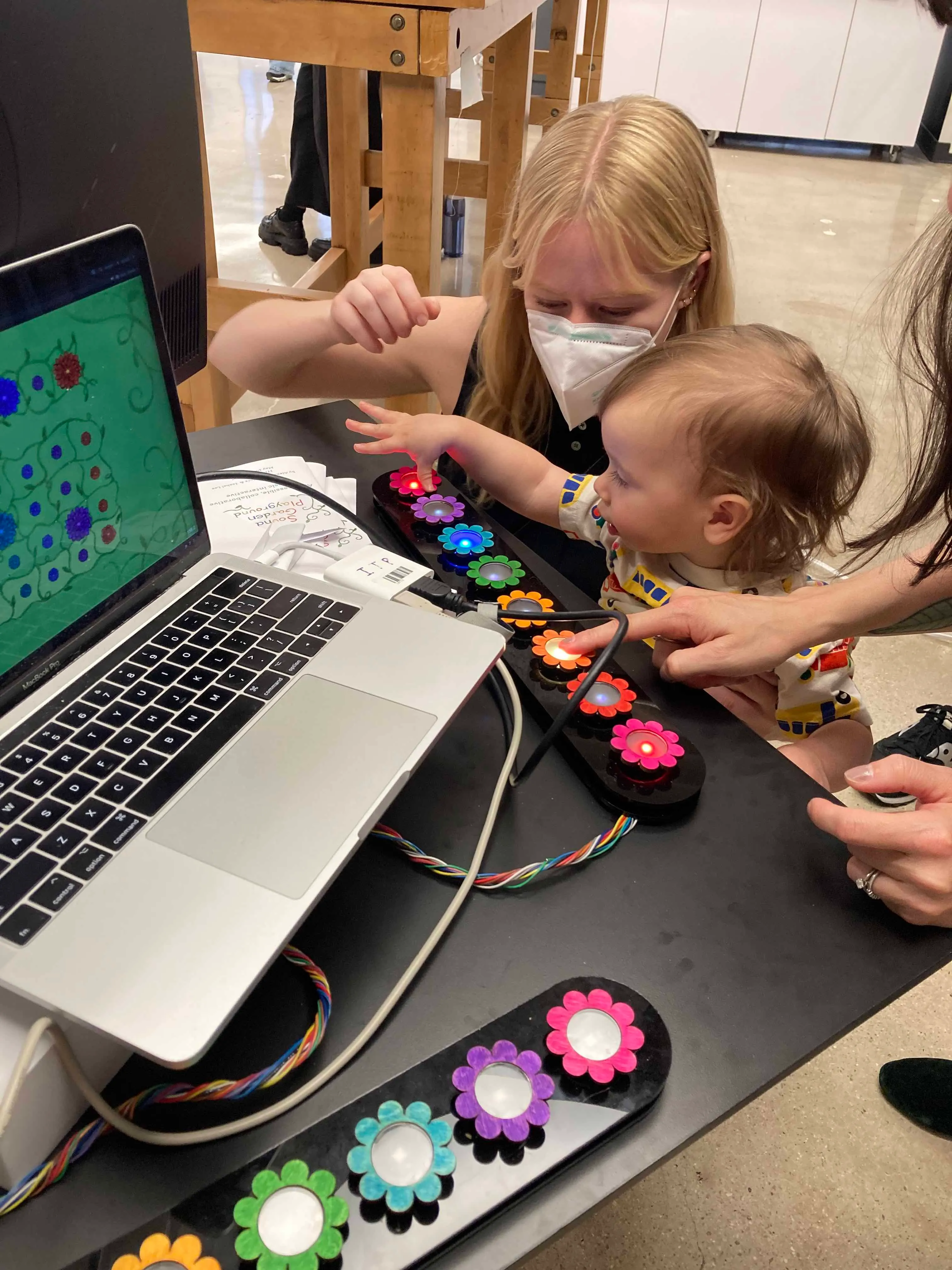 A mother and young child examine a row of buttons. The child reaches out to press one of the buttons that is lit up.