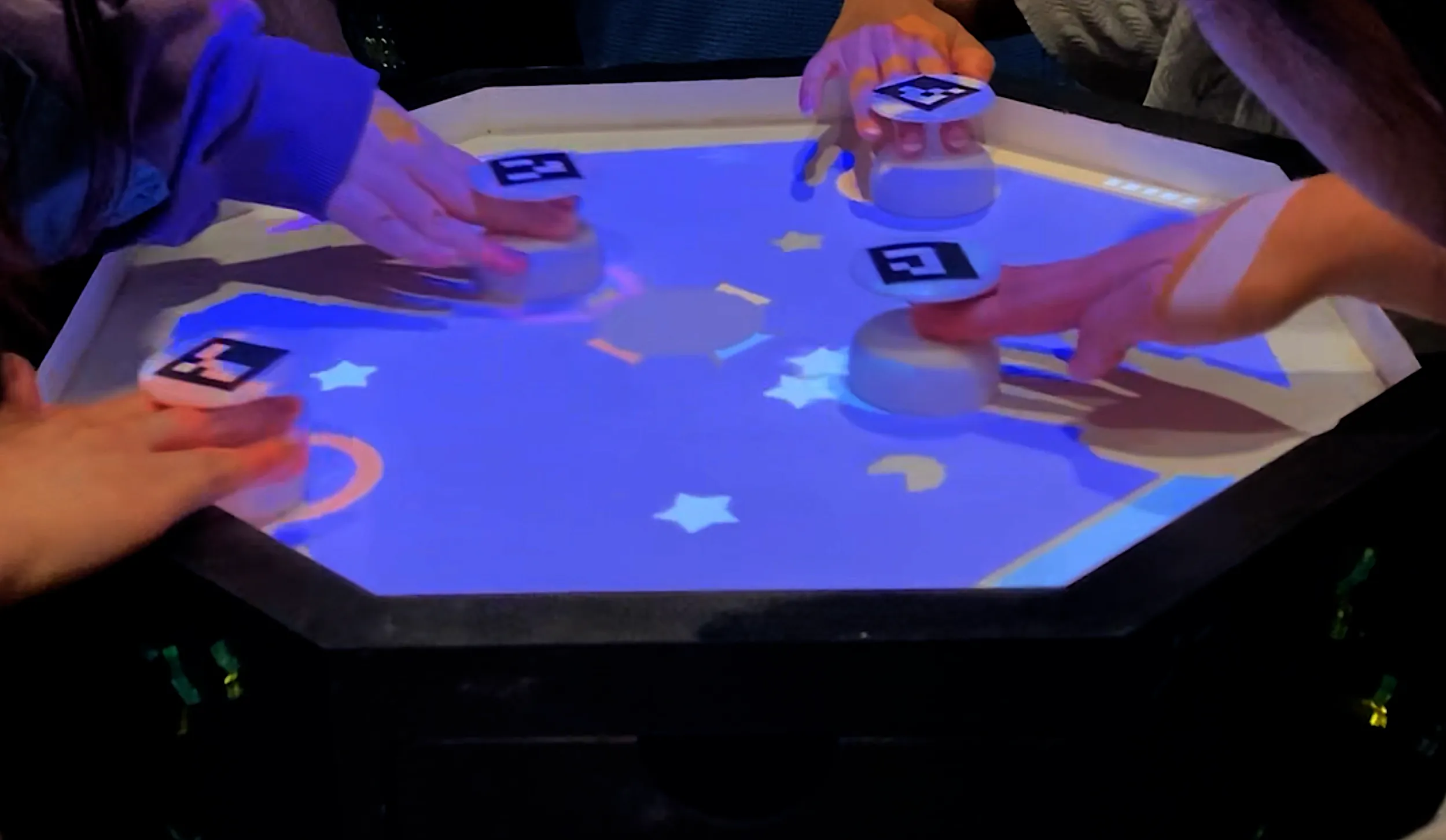 Four hands manipulate controllers in a projected playing space filled with stars. 
     In the foreground, a pac-man shape is projected alongside the stars. A red circle over the photo 
     indicates the position of the pac-man shape.