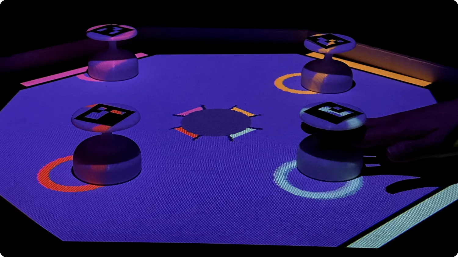 Four controllers sit atop a projected playing space on a table. Circles in different colors are 
    projected around each controller.