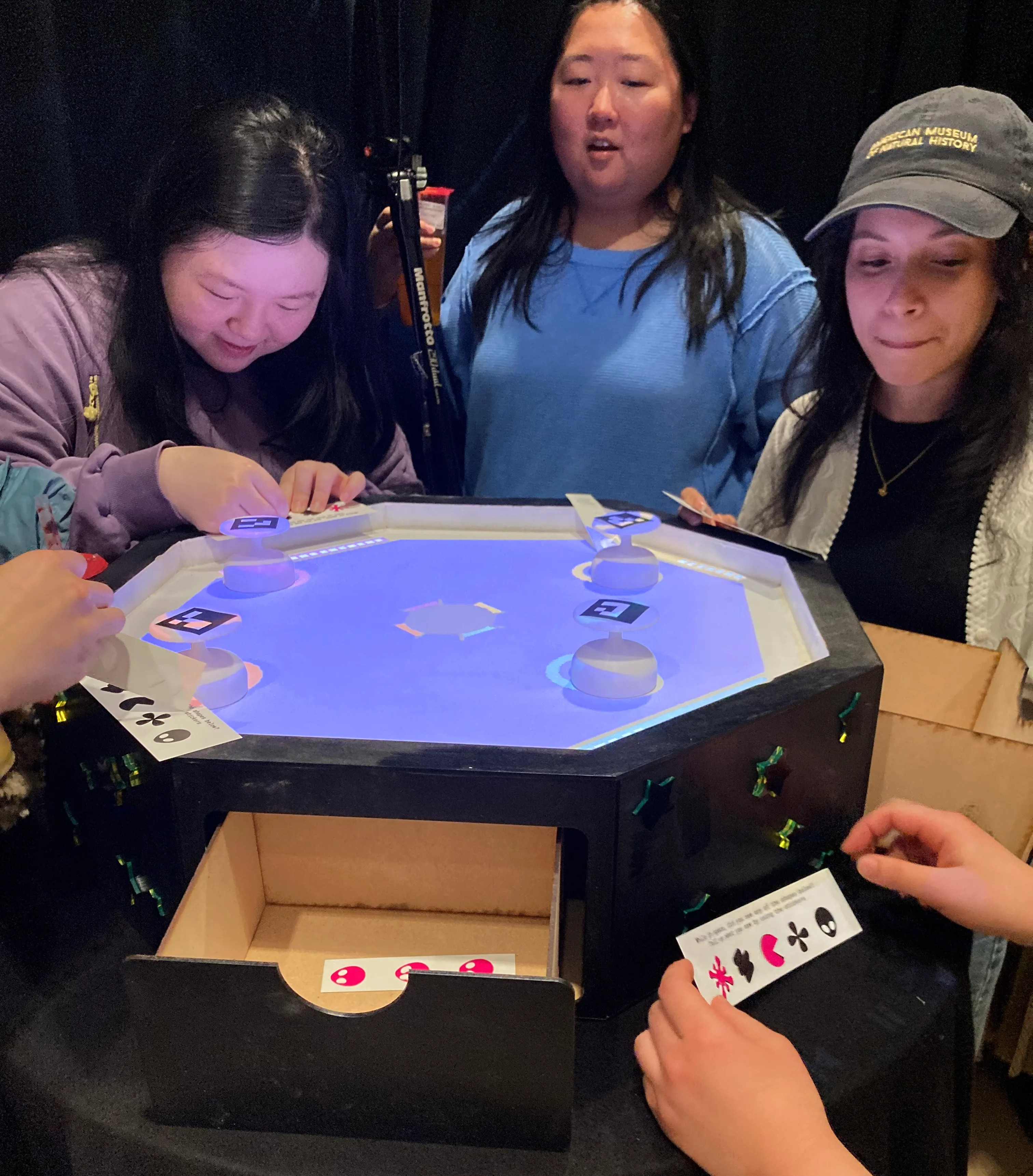 Five people stand around the game table, choosing shaped stickers to put on strips paper.
        A drawer in the side of the game table is open, showing a sheet of stickers inside.