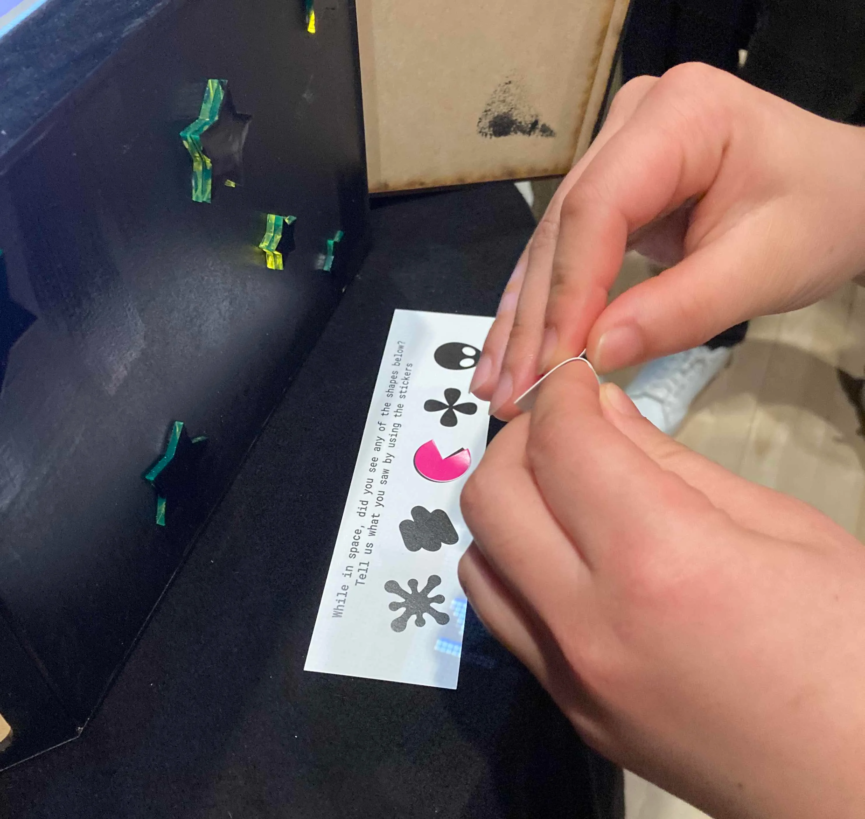 A pair of hands peel a sticker off a sheet. Under the hands, a strip of paper contains five black shapes 
    in a row: a snowflake, three lines, a pac-man, a four-petaled flower, and an alien head. The pac-man shape is 
    covered by a pink pac-man-shaped sticker. Text above the shapes reads 
    "While in space, did you see any of the shapes below? Tell us what you saw by using the stickers."